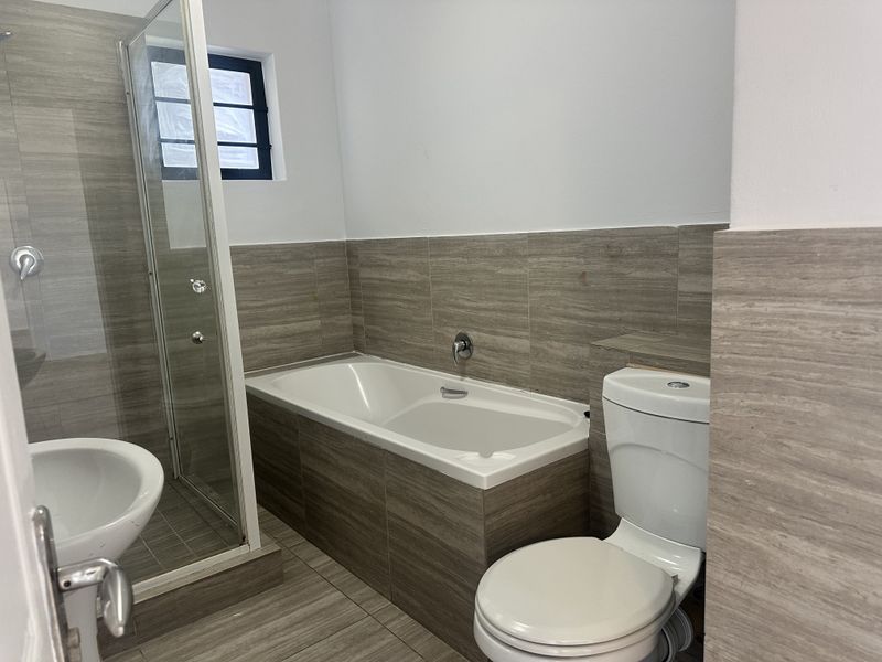 A modern bathroom featuring a built-in bathtub, a separate glass-enclosed shower, and a toilet, finished with neutral wood-look wall and floor tiles.