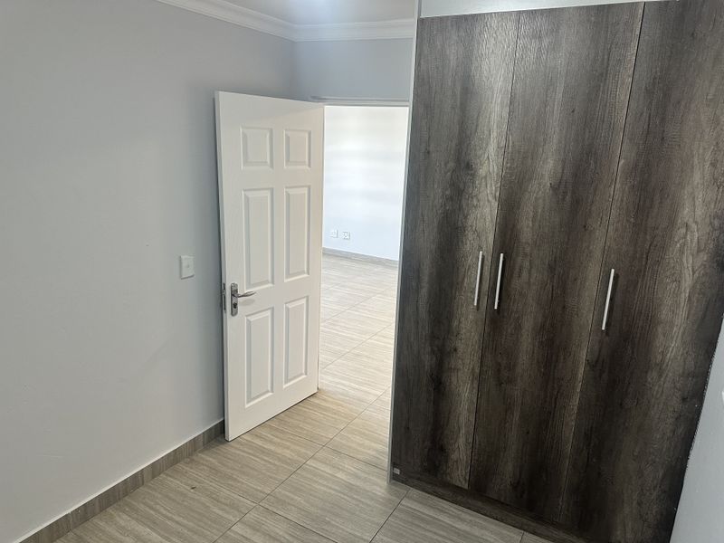 A clean, empty bedroom featuring modern wood-look built-in cupboards, neutral tiled flooring, and a white panel door leading to an adjacent room.