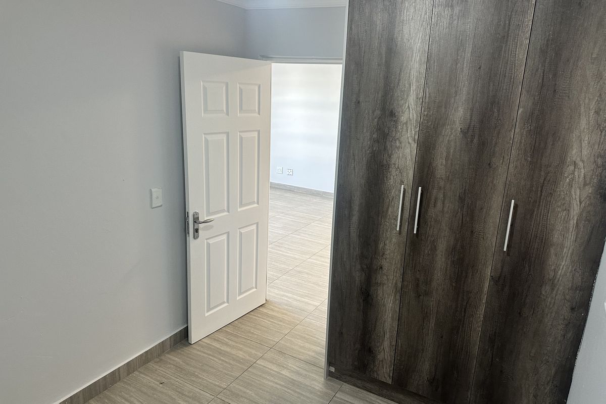 A clean, empty bedroom featuring modern wood-look built-in cupboards, neutral tiled flooring, and a white panel door leading to an adjacent room.
