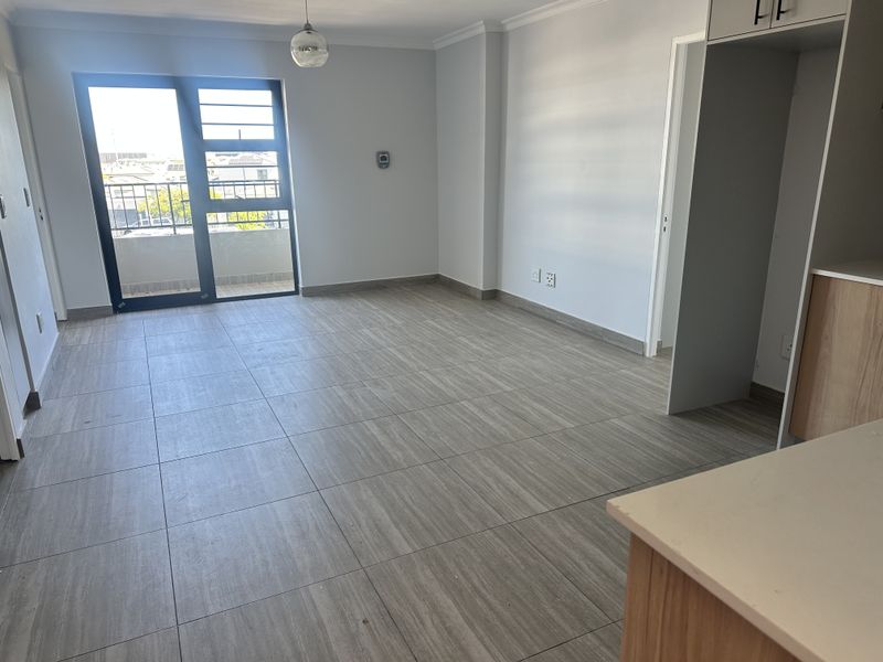 A bright, empty open-plan living and kitchen area featuring modern tiled flooring and a sliding glass door leading to a balcony.