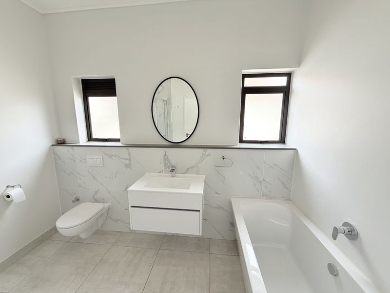 A modern, clean bathroom featuring a floating vanity, wall-mounted toilet, built-in bathtub, and elegant marble-look wall tiling with dual windows for natural light.