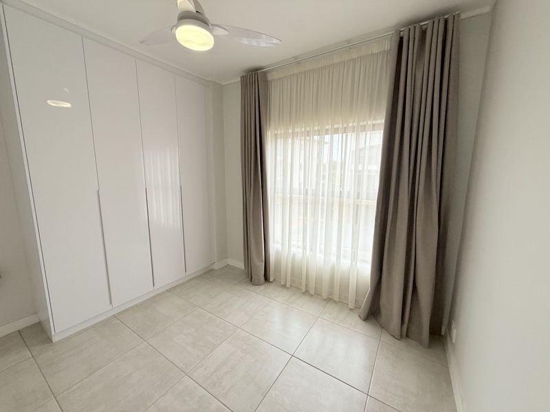 A bright, empty bedroom featuring modern white built-in cupboards, neutral tiled flooring, and a ceiling fan with integrated lighting.
