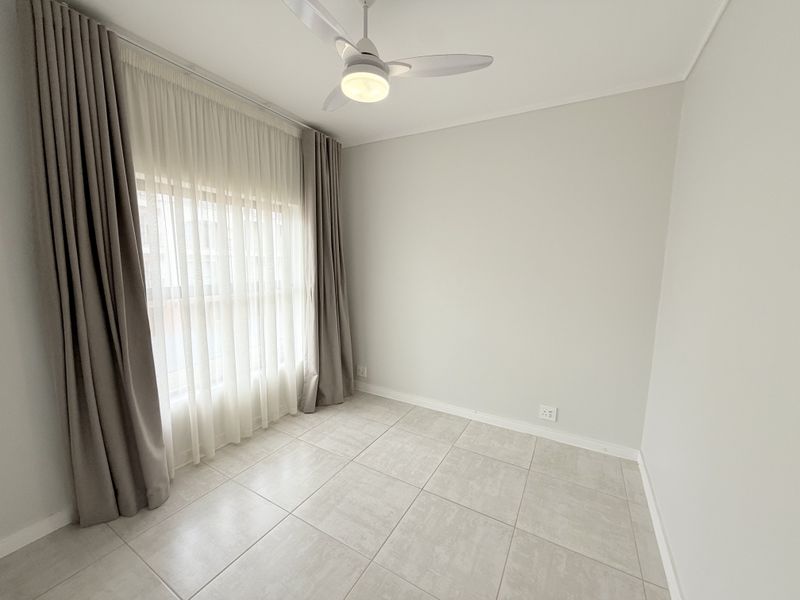 A bright, empty bedroom featuring neutral tiled flooring, a modern ceiling fan, and floor-to-ceiling curtains, offering a clean and versatile living space.