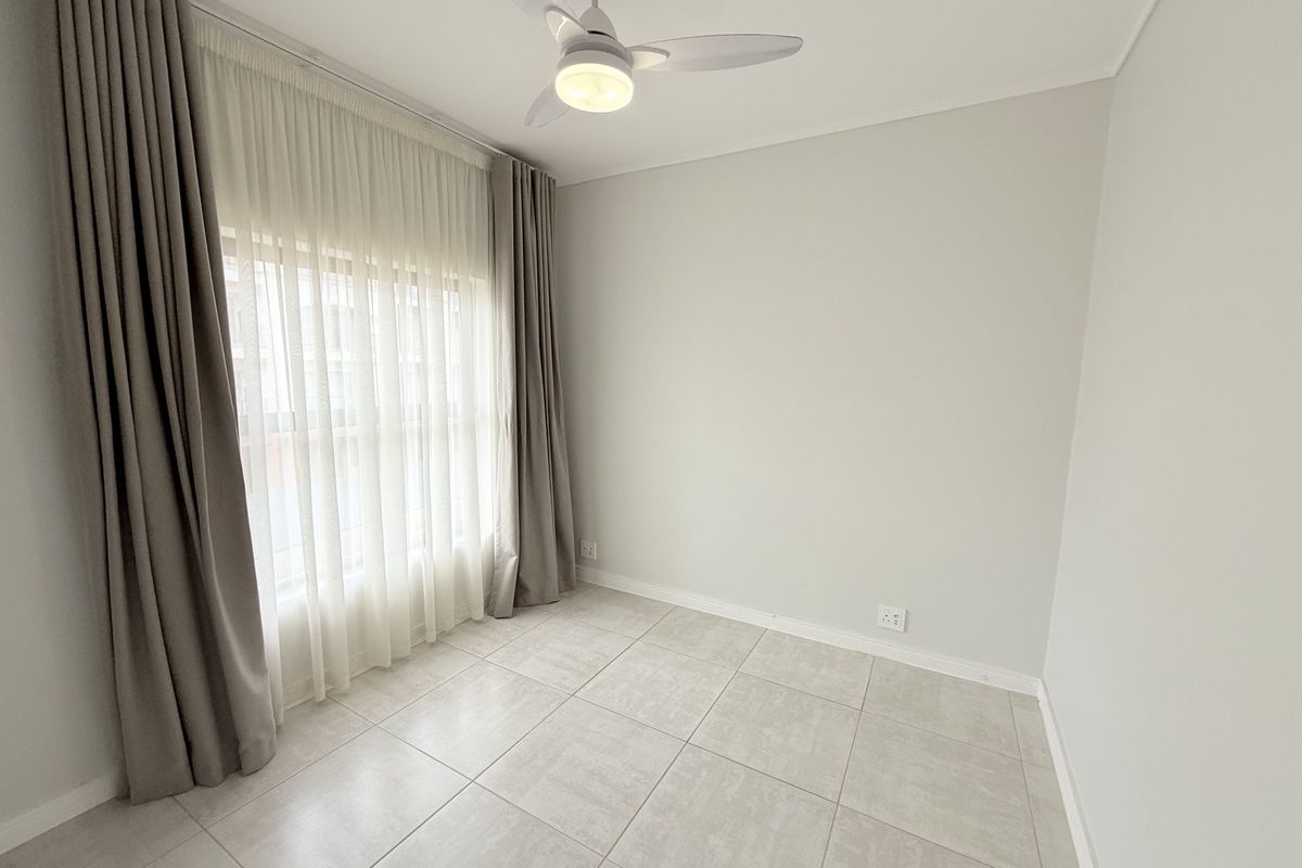 A bright, empty bedroom featuring neutral tiled flooring, a modern ceiling fan, and floor-to-ceiling curtains, offering a clean and versatile living space.
