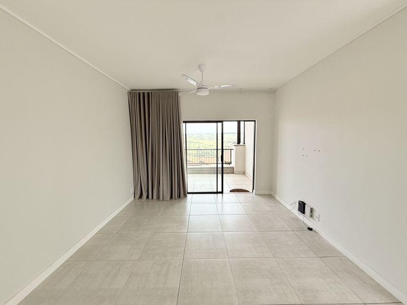 A bright, empty living room featuring neutral tiled flooring, a ceiling fan, and sliding glass doors leading to an outdoor balcony area.