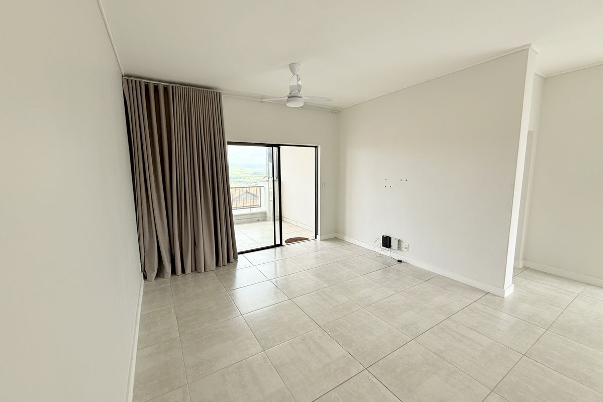 A bright, empty living room featuring neutral tiled flooring, a ceiling fan, and a sliding glass door providing access to an outdoor balcony.