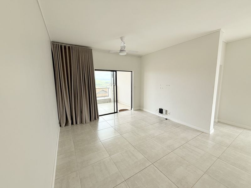 A bright, empty living room featuring neutral tiled flooring, a ceiling fan, and a sliding glass door providing access to an outdoor balcony.
