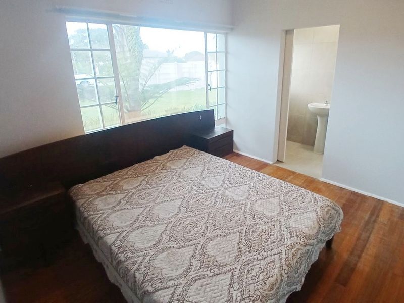 A bedroom featuring wooden flooring, a large window, and a built-in headboard with integrated bedside tables, leading into an en-suite bathroom.