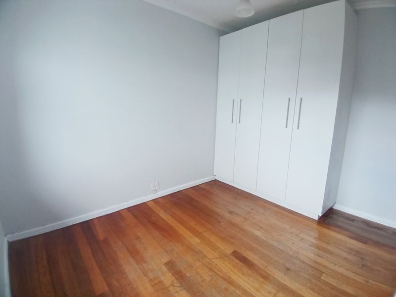 A bright, empty bedroom featuring polished hardwood flooring and a large, white built-in wardrobe with modern handles.