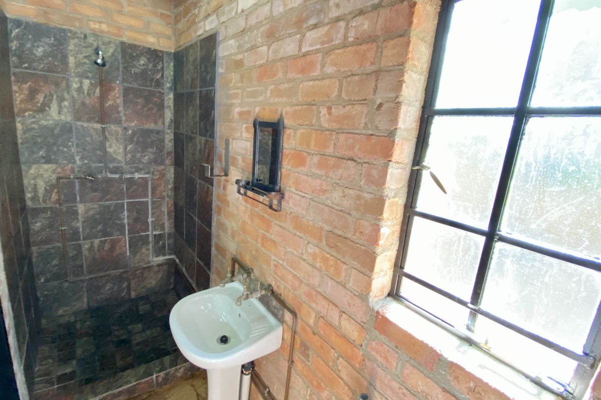 A rustic bathroom featuring exposed brick walls, a tiled shower enclosure, pedestal basin, and steel-framed window with industrial-style exposed copper plumbing.