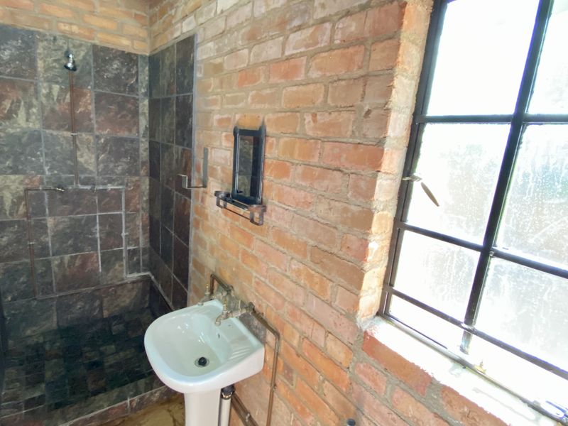 A rustic bathroom featuring exposed brick walls, a tiled shower enclosure, pedestal basin, and steel-framed window with industrial-style exposed copper plumbing.