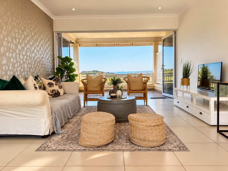 Bright living room featuring large bi-fold doors that open onto a covered patio with scenic views, complemented by neutral tiled flooring and textured wallpaper.