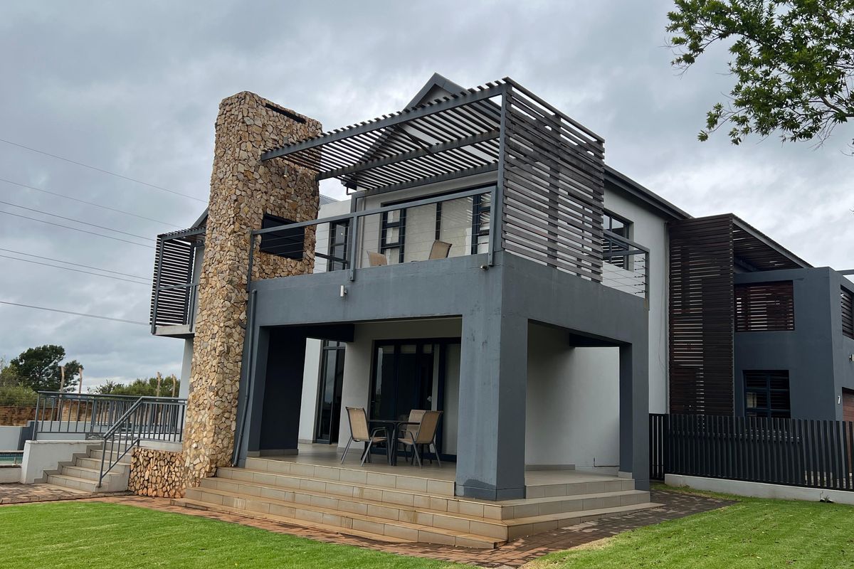 Rear view of a modern double-story house featuring a stone-clad chimney, a covered patio, and an upper-level balcony with timber pergola detailing.