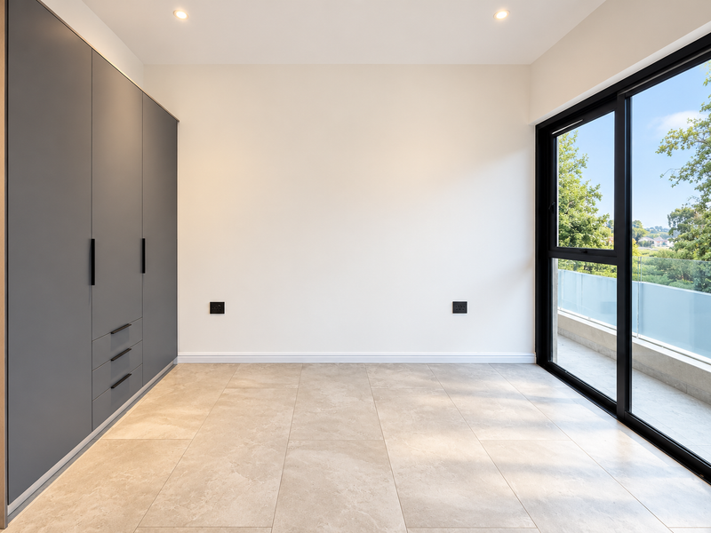 A bright, modern bedroom featuring sleek grey built-in cupboards, large format floor tiles, and a sliding door leading to a balcony with glass balustrades.