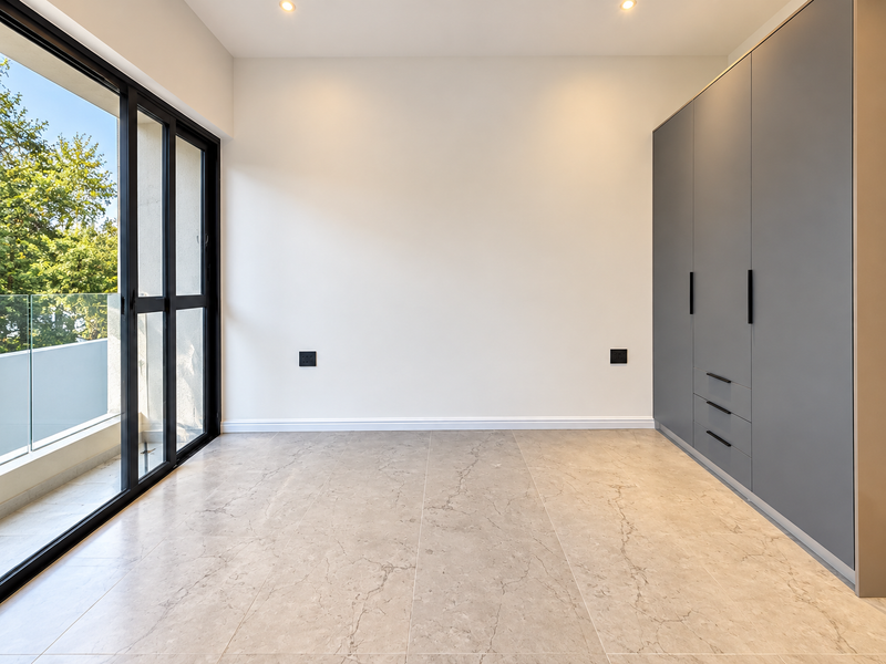 A bright, modern bedroom featuring sleek grey built-in cupboards, large sliding glass doors leading to a balcony, and high-quality tiled flooring.