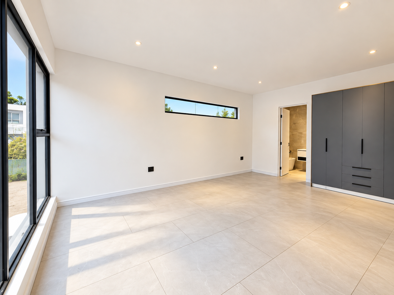 A modern, spacious bedroom featuring large windows, neutral tiled flooring, recessed lighting, and sleek built-in cupboards with an en-suite bathroom access.