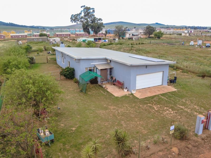 Aerial view of a detached single-story house featuring a metal roof with solar panels, a garage, and a large surrounding garden plot.