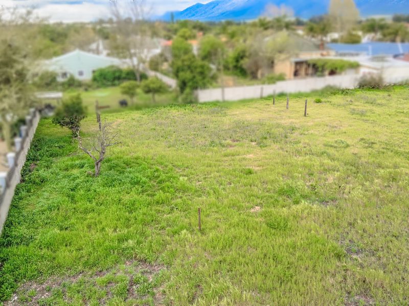 An elevated aerial view of a vacant residential plot featuring grass coverage, boundary walls, and visible survey pegs.