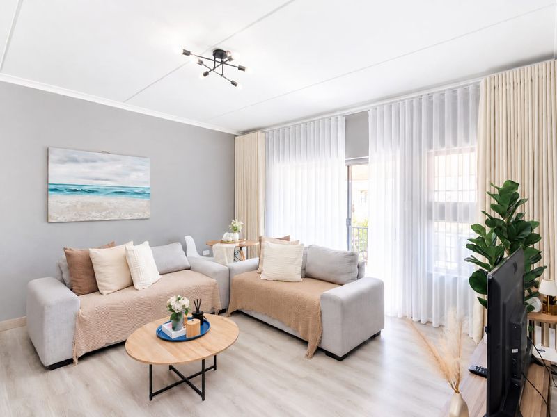 A bright and well-maintained living room featuring light laminate flooring, modern ceiling lighting, and large sliding glass doors with floor-to-ceiling curtains.