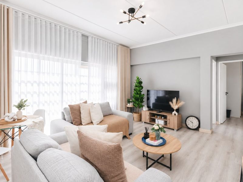 A bright and well-maintained living room featuring light-toned laminate flooring, neutral wall finishes, and large windows with floor-to-ceiling curtains.