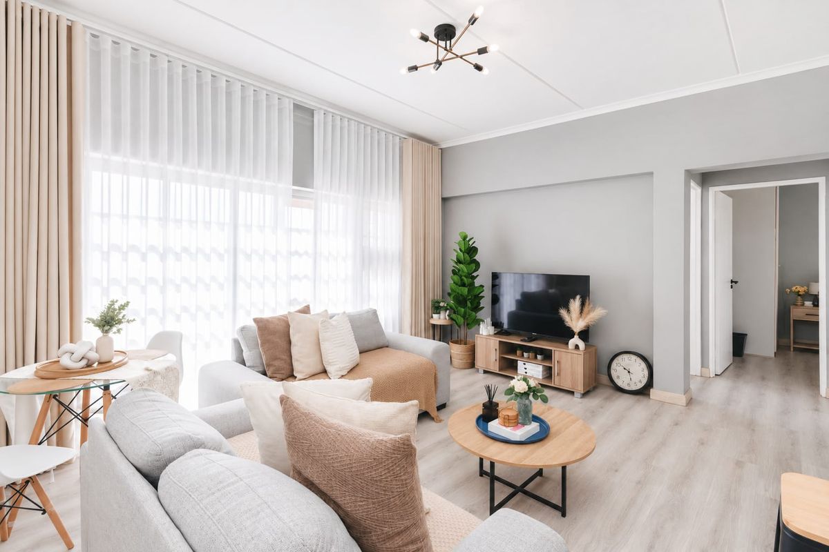 A bright and well-maintained living room featuring light-toned laminate flooring, neutral wall finishes, and large windows with floor-to-ceiling curtains.