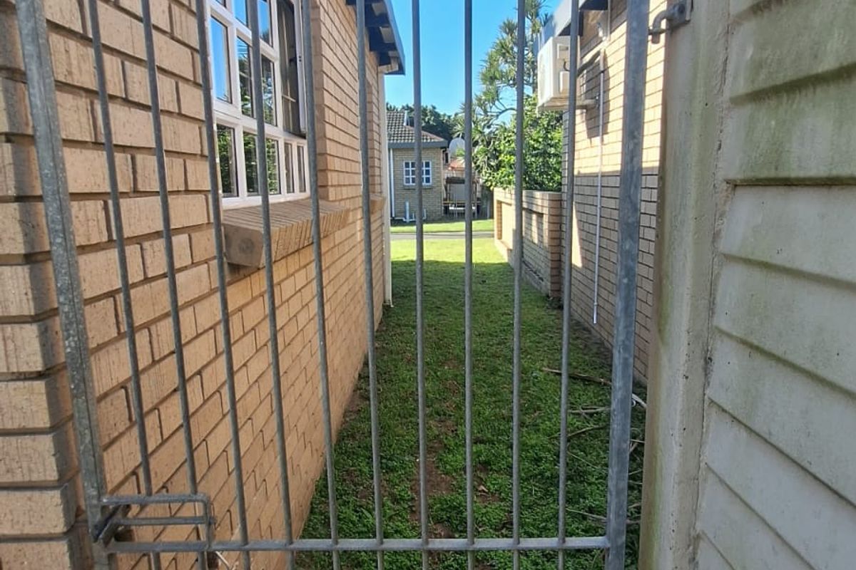 A narrow side passage between two townhouse units featuring a metal security gate, face brick exterior, and a small grassed area.