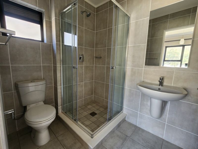 A clean, modern bathroom featuring a glass-enclosed shower, pedestal basin, wall-mounted mirror, and neutral floor-to-ceiling tiling.