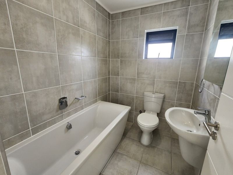 A clean, modern bathroom featuring neutral grey wall and floor tiles, a white bathtub, pedestal sink, and toilet.