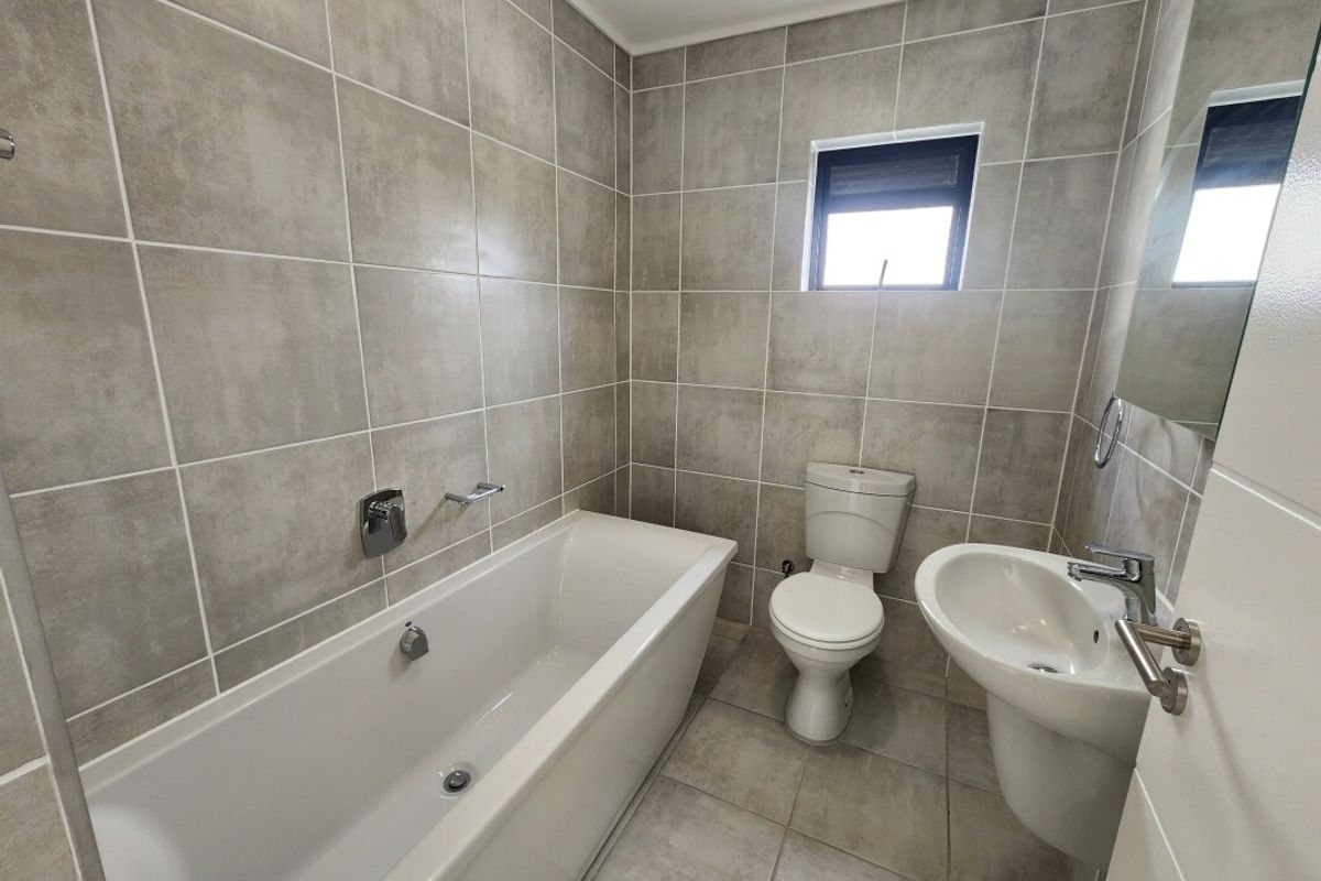 A clean, modern bathroom featuring neutral grey wall and floor tiles, a white bathtub, pedestal sink, and toilet.