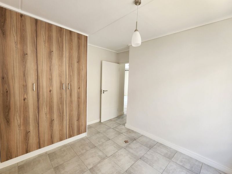 A bright, empty bedroom featuring modern wood-finish built-in cupboards, neutral wall paint, and light grey floor tiles.