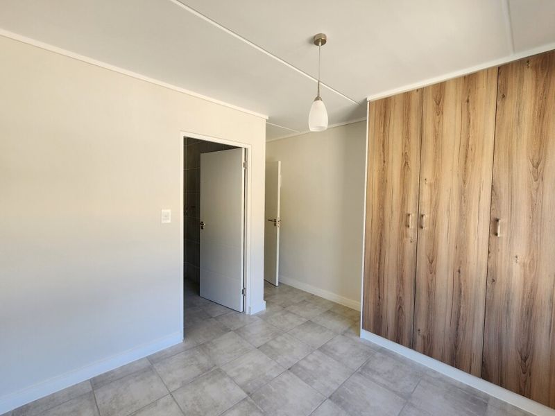 A clean, empty bedroom featuring modern wood-finish built-in cupboards, neutral tiled flooring, and a simple pendant light fixture.