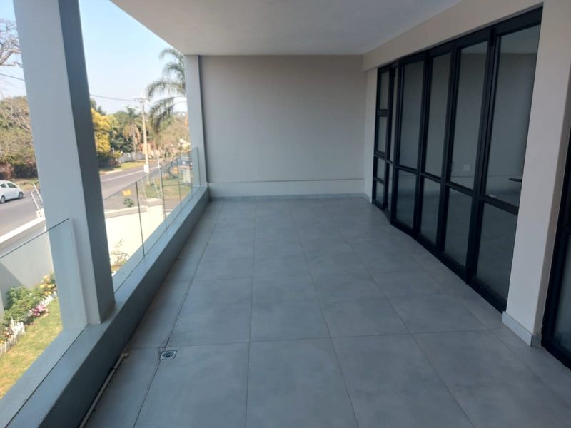 A spacious, modern covered balcony featuring large-format floor tiles, a sleek glass balustrade, and black aluminium framed sliding doors leading to the interior.