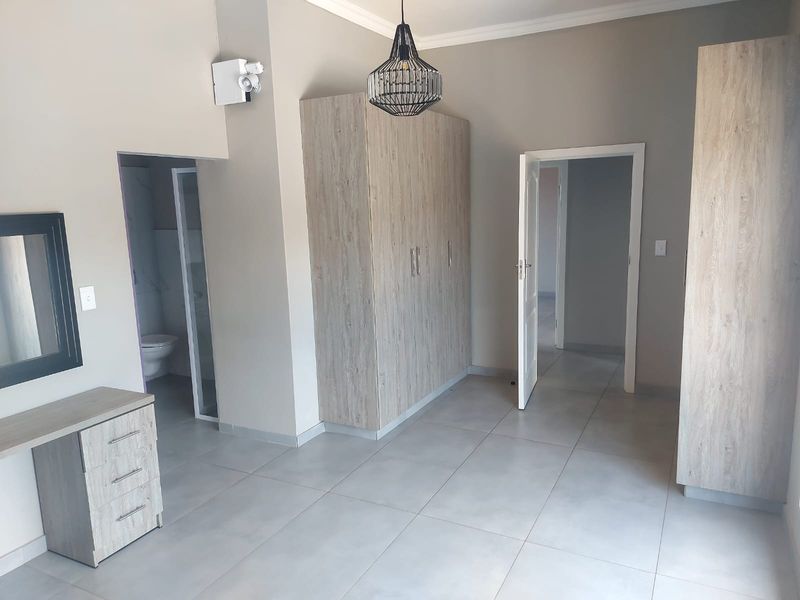 Spacious bedroom featuring neutral tiled flooring, built-in cupboards, and an en-suite bathroom, offering a clean and modern living space.