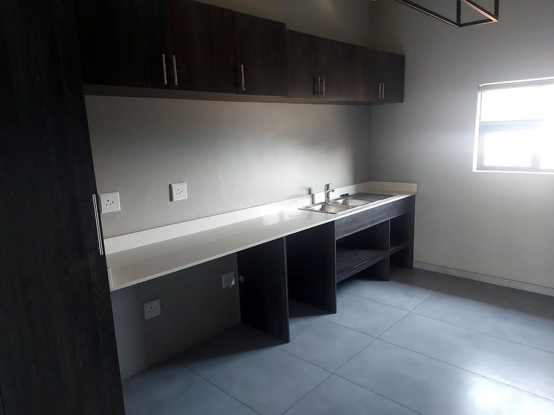 A modern, empty kitchen featuring dark wood cabinetry, stone countertops, a stainless steel sink, and neutral tiled flooring.