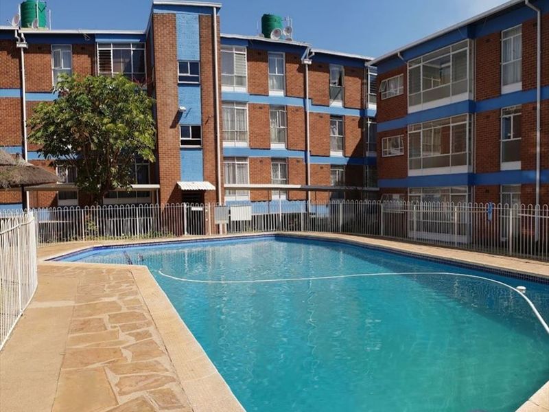 Communal swimming pool area featuring paved surrounds and metal perimeter fencing, set against a multi-story brick apartment building.