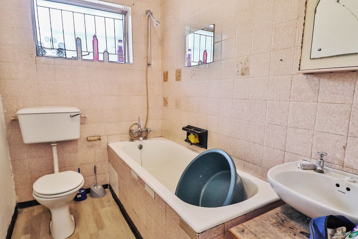 A dated bathroom featuring a bathtub, pedestal basin, and toilet, with tiled walls and security bars on the window.