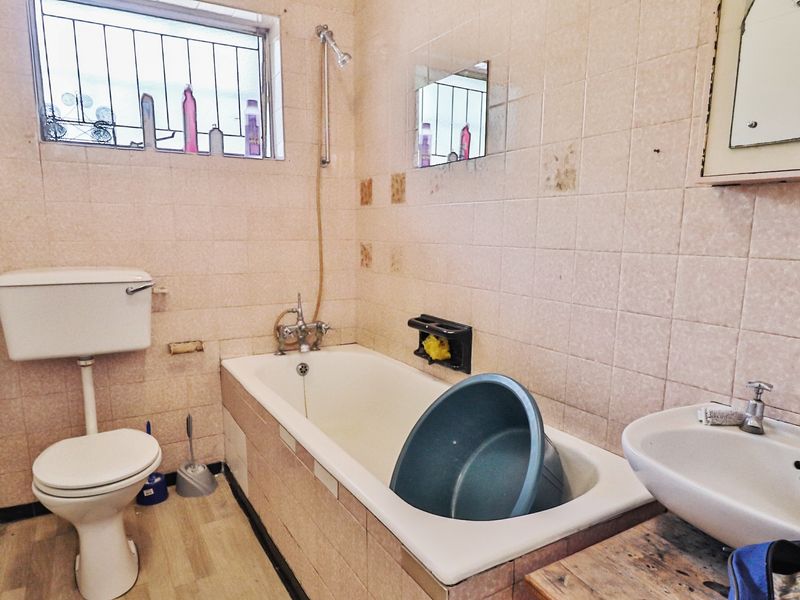 A dated bathroom featuring a bathtub, pedestal basin, and toilet, with tiled walls and security bars on the window.