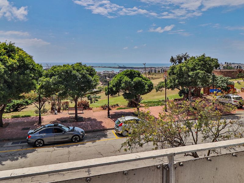 Elevated view overlooking a landscaped park, street, and distant coastal horizon from a balcony with a metal balustrade.