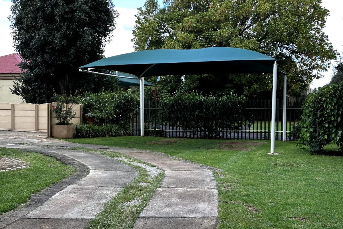 A well-maintained garden area featuring a green shade sail structure, concrete driveway, and secure perimeter fencing.