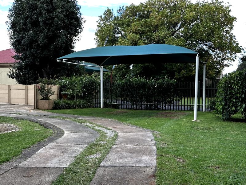 A well-maintained garden area featuring a green shade sail structure, concrete driveway, and secure perimeter fencing.