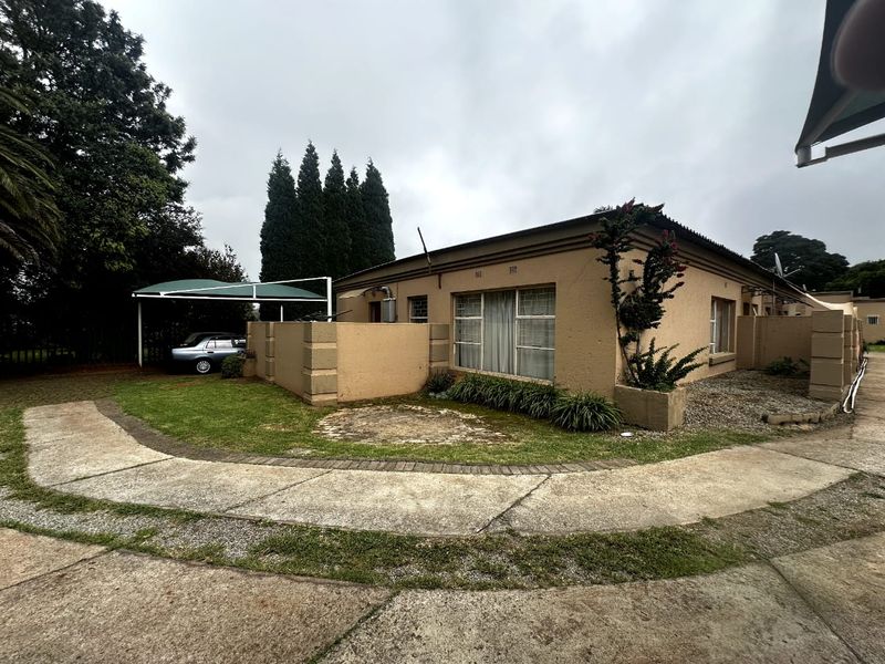Front exterior view of a single-story house featuring a concrete driveway, carport, and boundary wall with established garden beds.