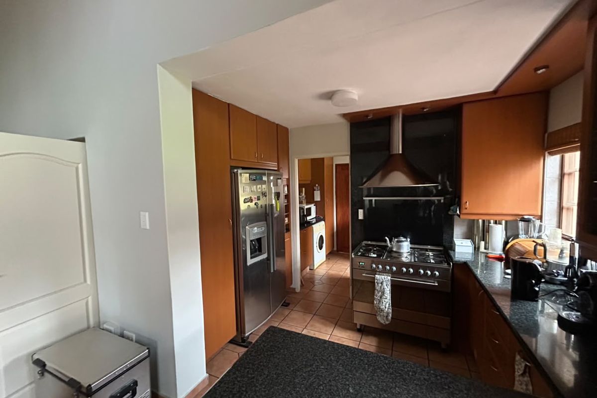 A functional kitchen featuring wooden cabinetry, granite countertops, a stainless steel gas stove with extractor fan, and tiled flooring, with an adjacent scullery area.