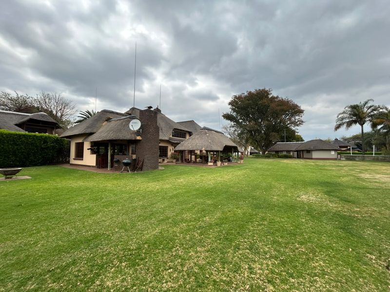 Spacious rear garden featuring a large lawn, a main house with a traditional thatch roof, and a separate covered outdoor entertainment area.