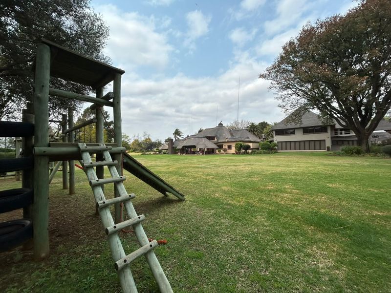 A spacious rear garden featuring a large lawn, mature trees, and a timber play structure, with views of the thatched-roof main residence.