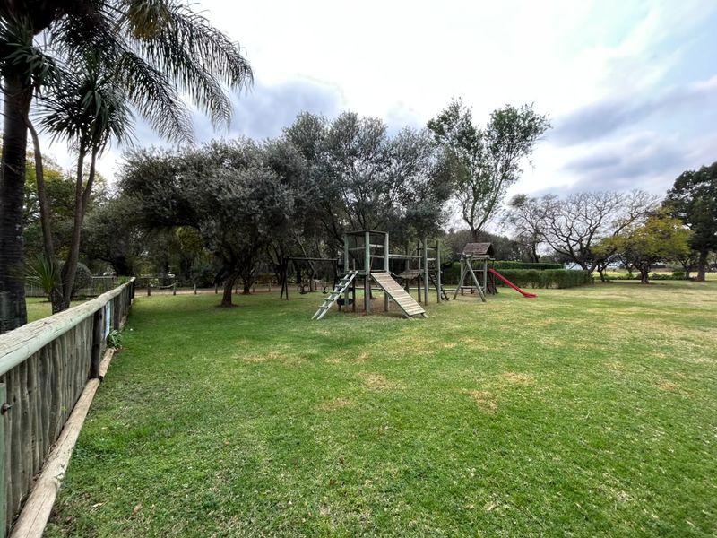 A spacious, well-maintained garden featuring mature trees, a large lawn area, and a wooden playground structure enclosed by a timber fence.