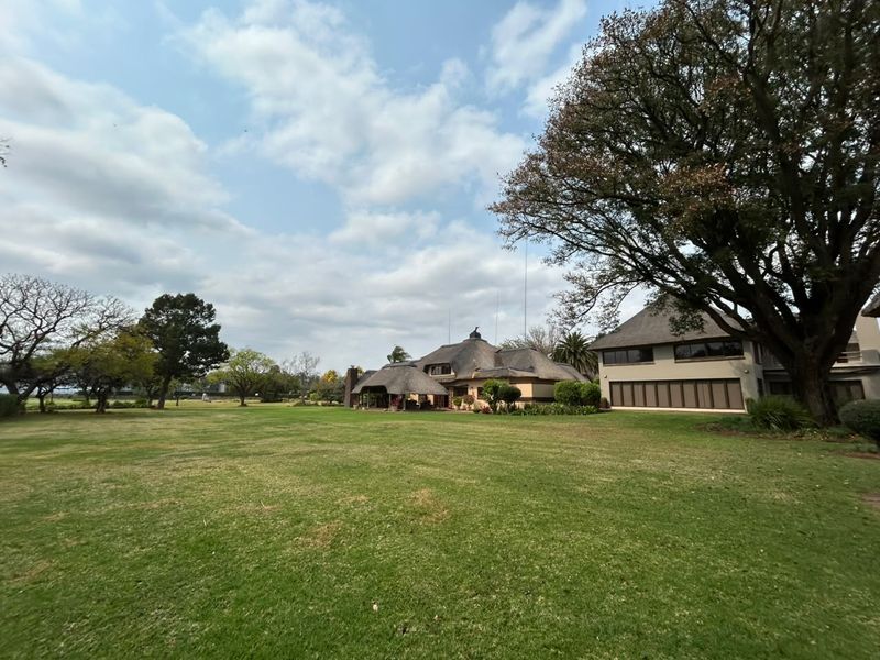 A wide-angle view of the rear elevation featuring a large landscaped garden, mature trees, and a traditional thatched roof residence with a covered patio area.
