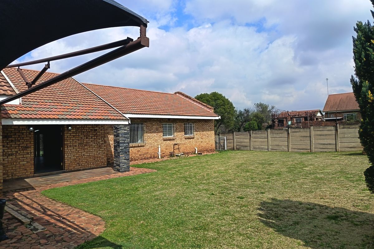 Rear view of the house featuring a brick facade, tiled roof, paved patio area with a shade sail, and a spacious lawn with boundary wall.