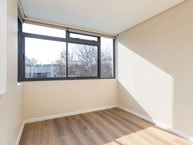 A bright, empty bedroom featuring modern laminate wood flooring, neutral walls, and large aluminium-framed windows providing natural light and an urban view.