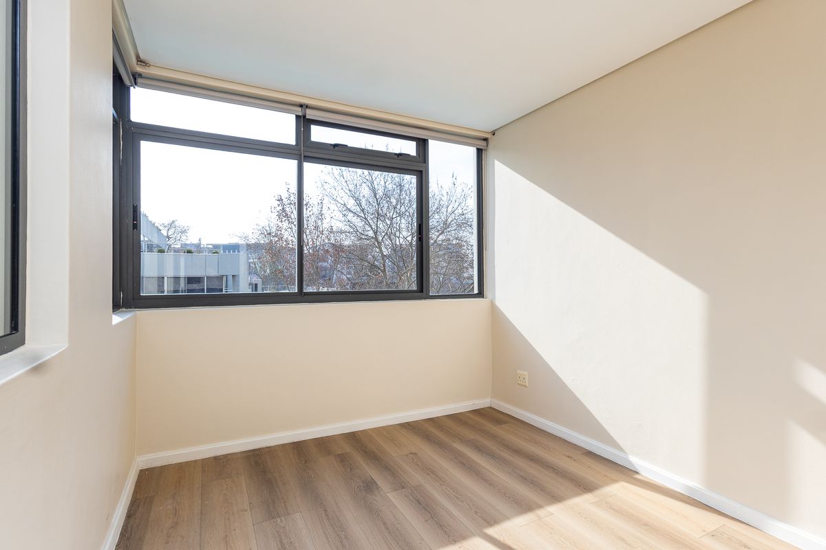 A bright, empty bedroom featuring modern laminate wood flooring, neutral walls, and large aluminium-framed windows providing natural light and an urban view.