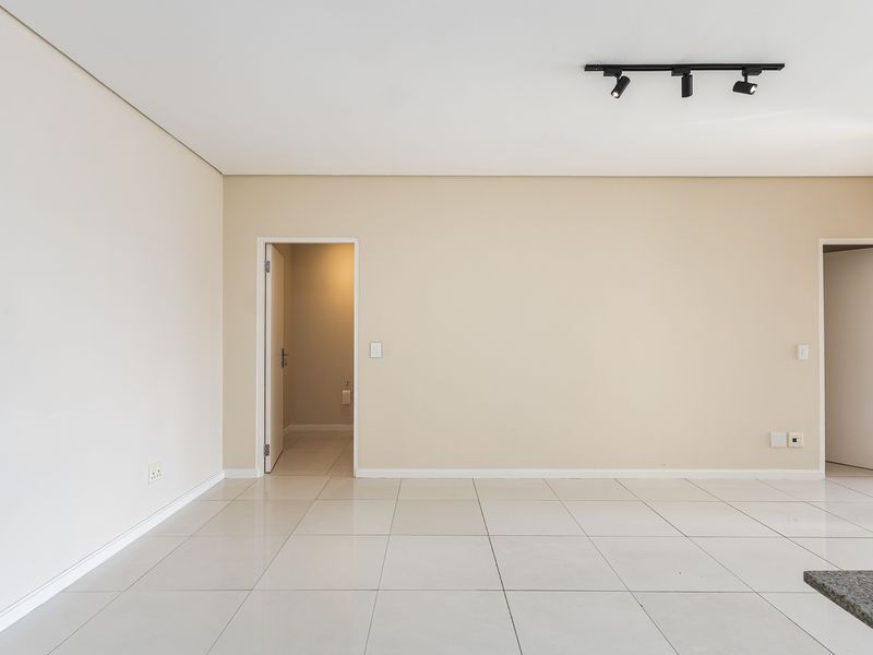 A bright, empty living area featuring neutral walls, large format floor tiles, and modern track lighting, providing a clean canvas for interior design.